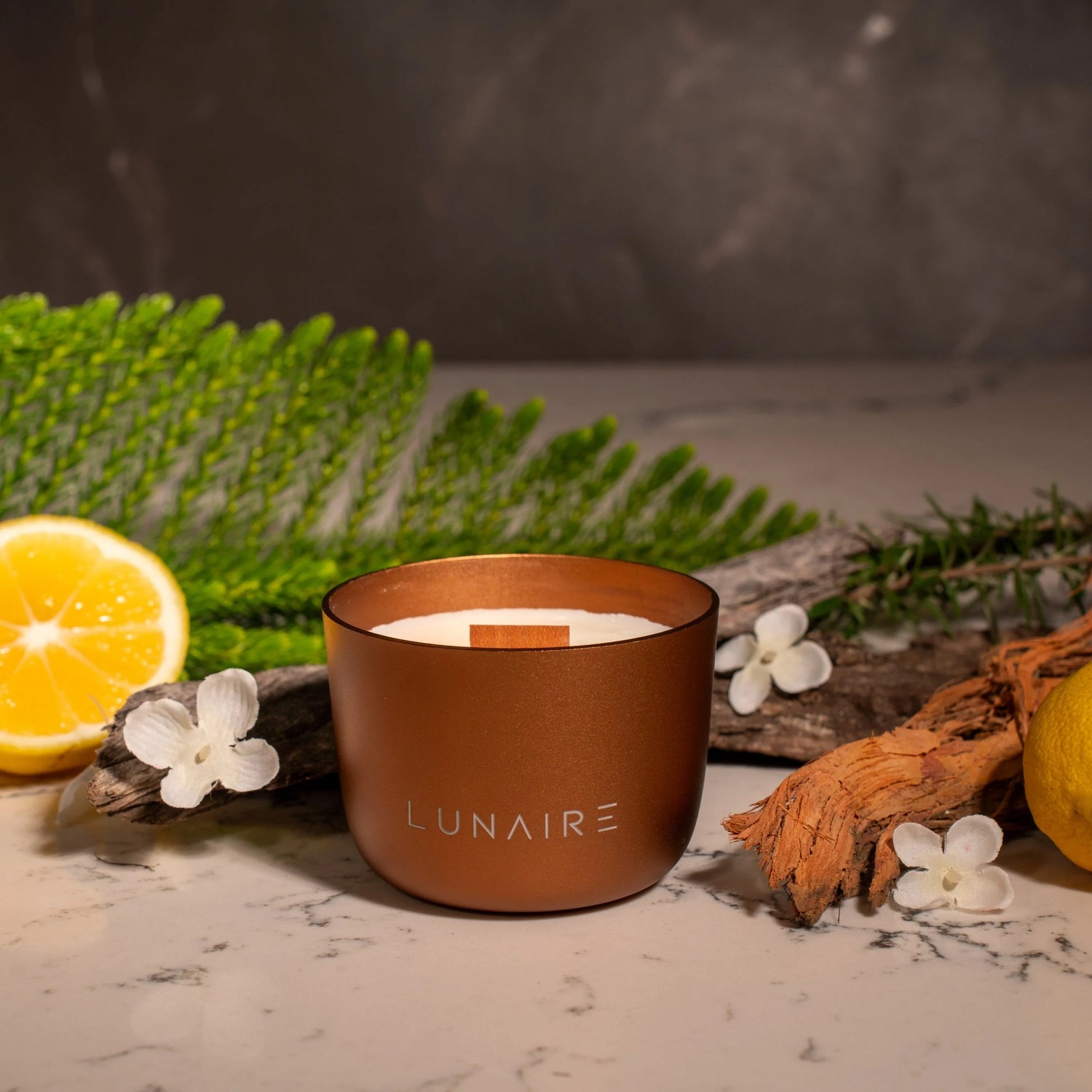 Bronze candle with 'LUNAIRE' branding on a marble surface with lemon, flowers, and greenery.