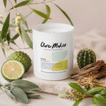 A white glass jar candle labeled 'Clare Makes' sits on a stone surface surrounded by botanical props including eucalyptus leaves, a lime, a cactus, sage, dried bark, and a small dried flower sprig. The candle is lit with a warm glow, and the background is a soft beige fabric.