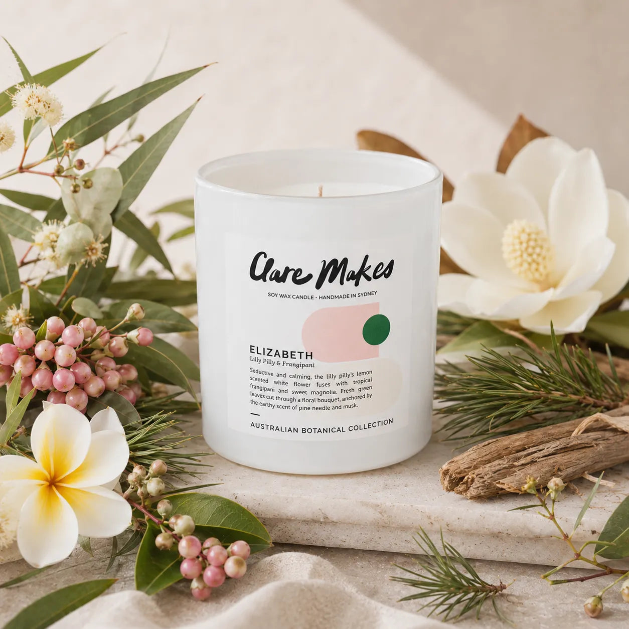 A white glass candle jar labeled 'Clare Makes Elizabeth' styled on a stone surface with white flowers, green leaves, pink berries, pine needles, and driftwood.