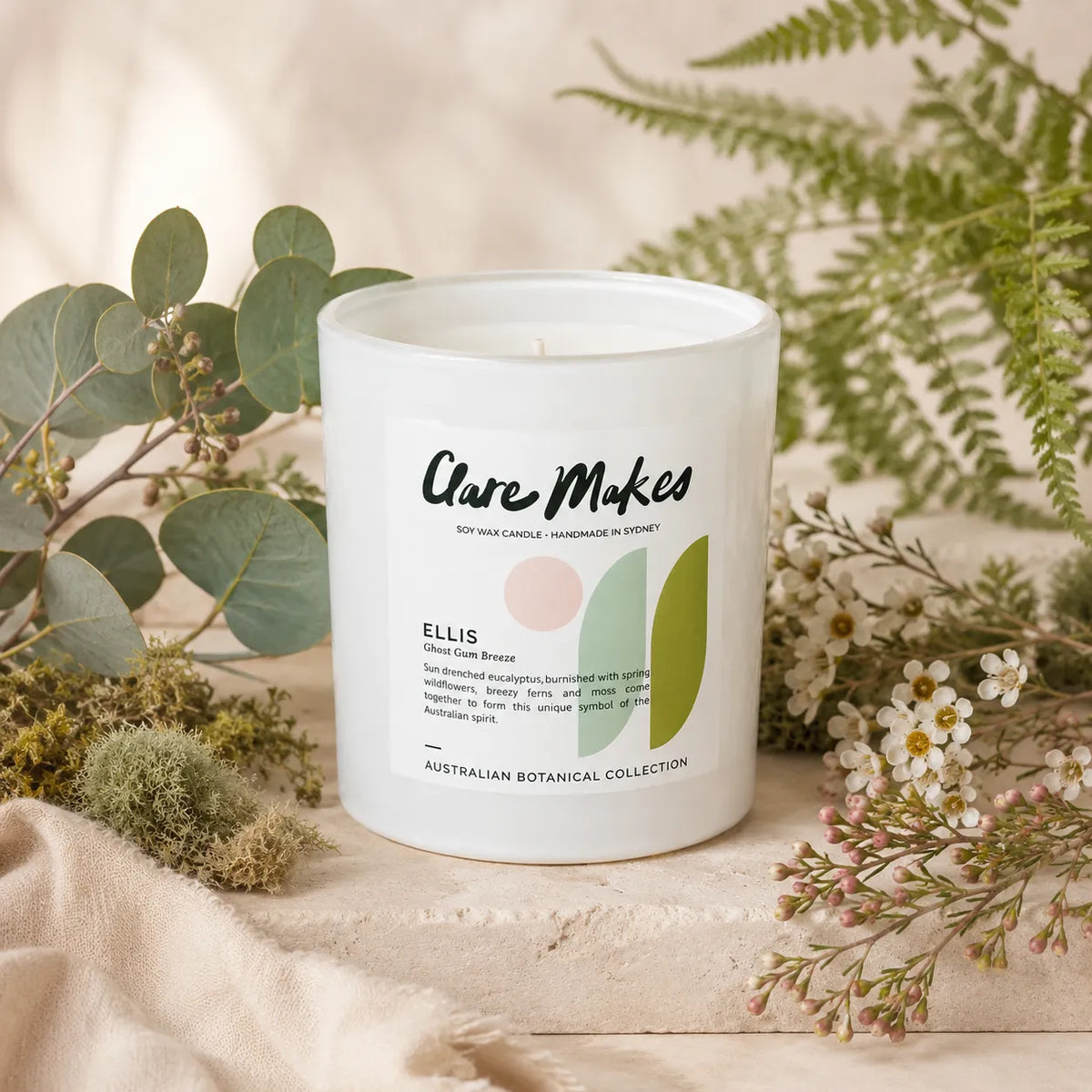A white glass jar candle labeled 'Care Makes' and 'Ellis' from the Australian Botanical Collection, styled on a beige fabric surface with green eucalyptus and white flowers.