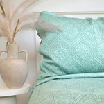 Teal patterned comforter on a bed with a vase and decorative feathers in the background.