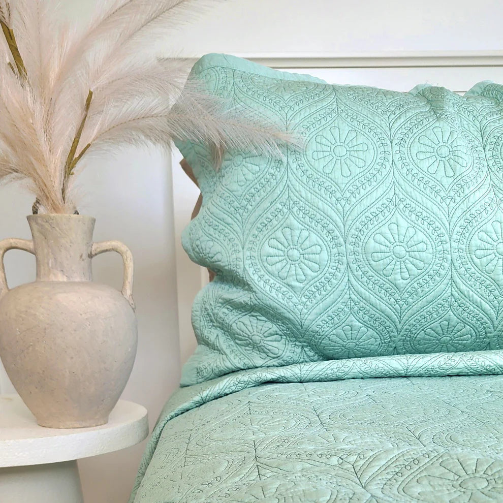 Teal patterned comforter on a bed with a vase and decorative feathers in the background.