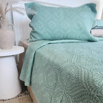 Teal patterned bedspread on a bed with a white side table and decorative plant.