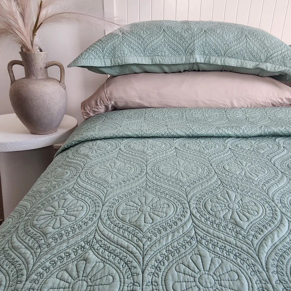 Teal patterned bedding set on a bed with a side table and vase in the background.