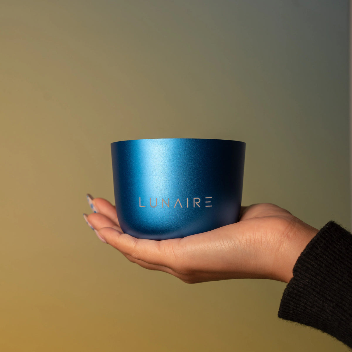 Hand holding a blue cylindrical container with 'LUNAIRE' branding against a gradient background