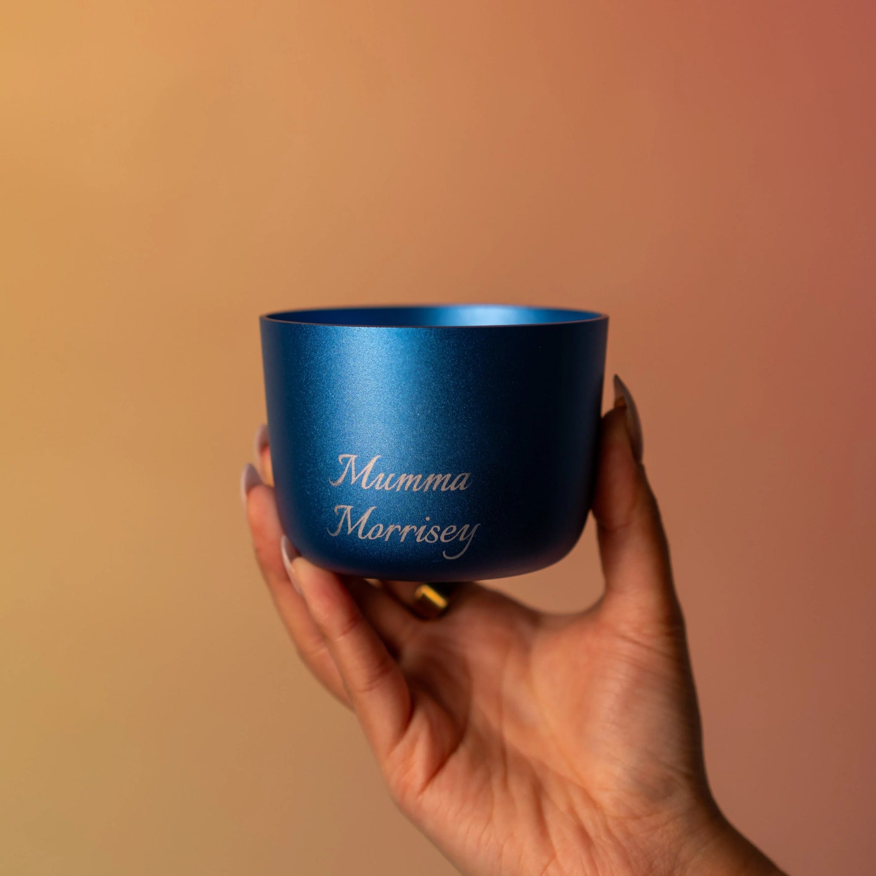 Hand holding a blue cup with 'Mumma Morrisey' text against a warm gradient background