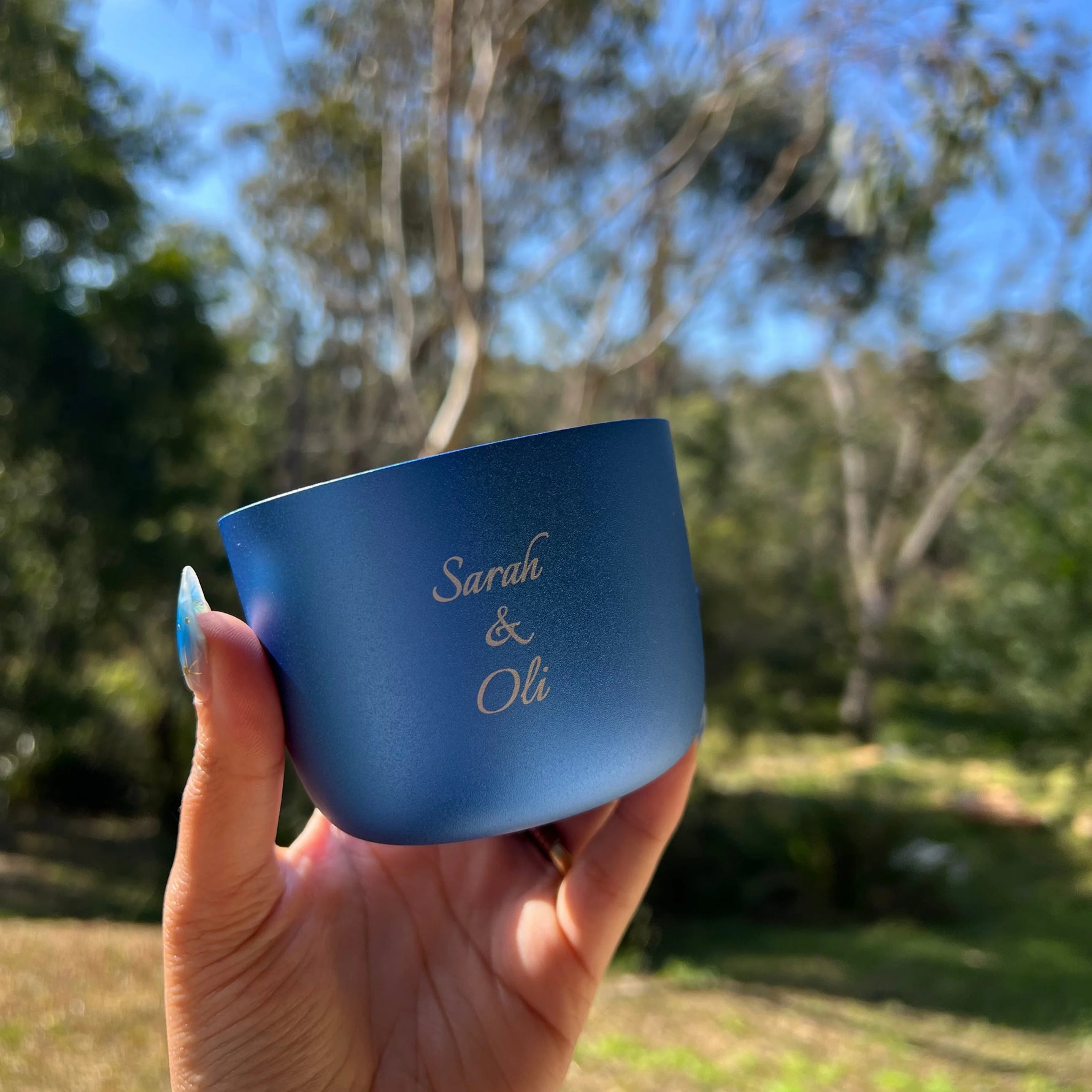 Hand holding a blue mug with 'Sarah & Oli' engraving against a blurred natural background
