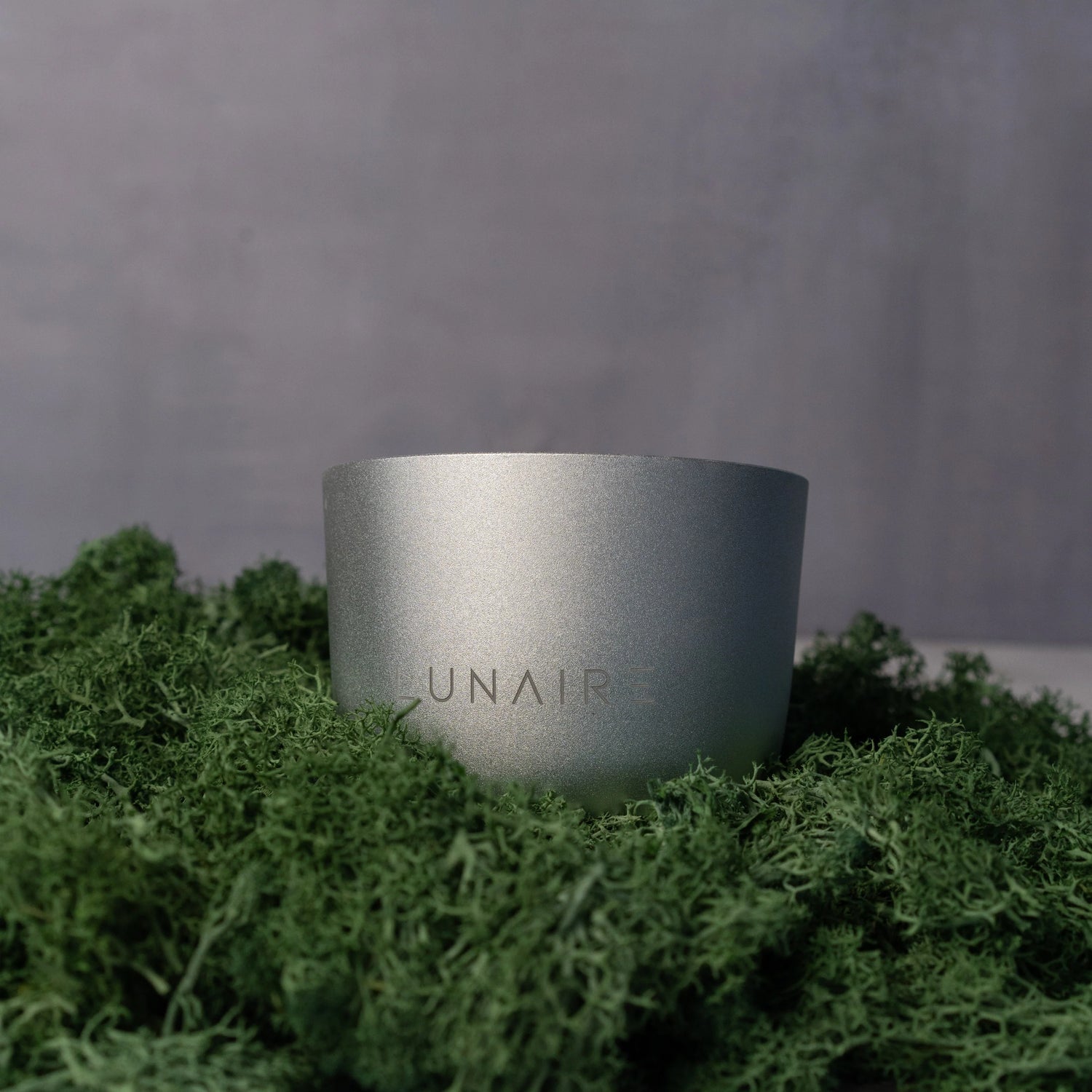 Silver cylindrical container with 'LUNAIR' branding on a green moss background