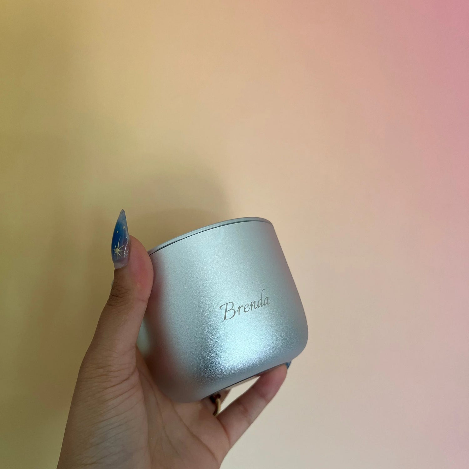 Hand holding a silver container with 'Brenda' engraved on it against a blurred background