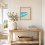 A framed square art print of a pastel beach and ocean scene hangs on a white paneled wall above a light wood console table styled with a vase of greenery, books, and a woven basket.