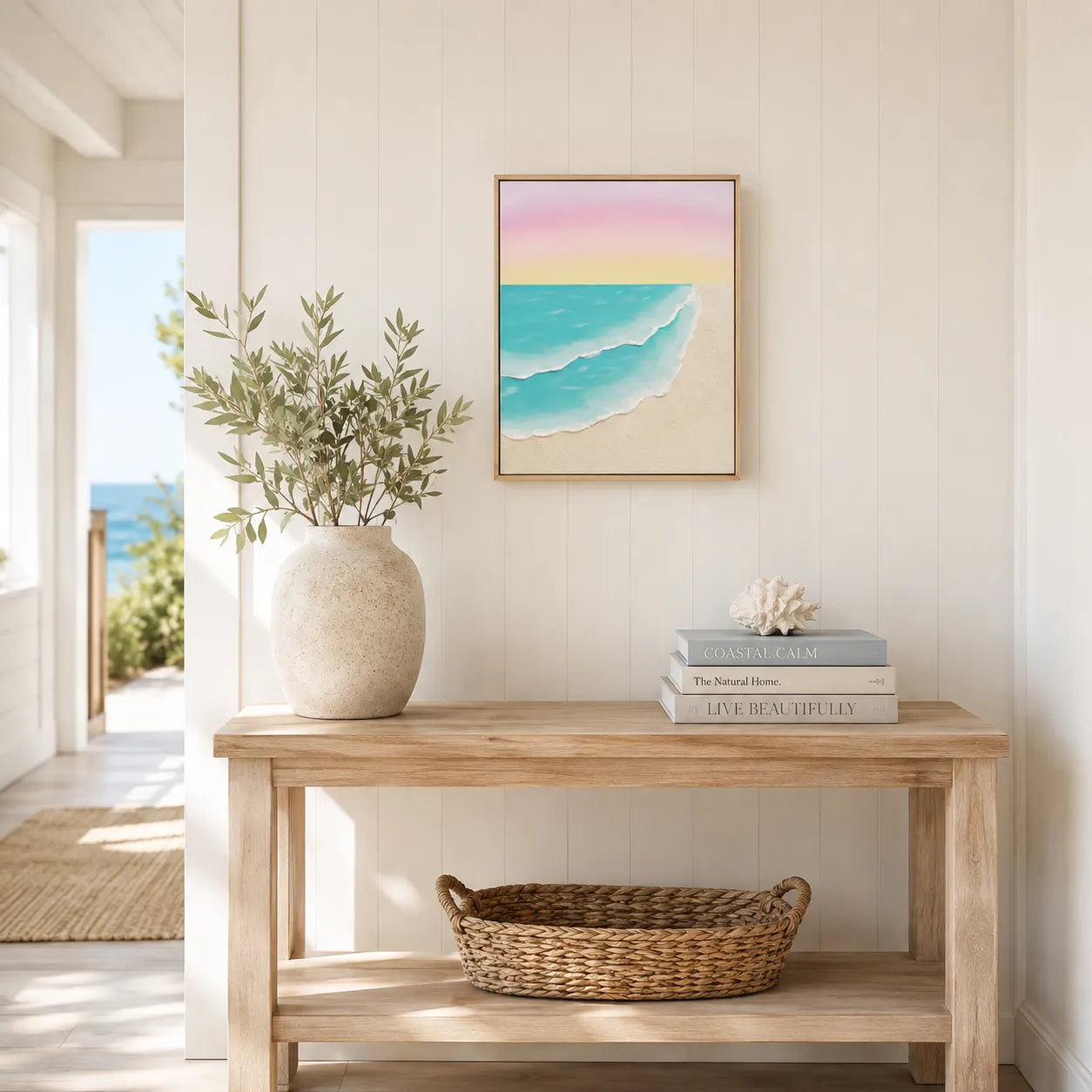 A framed square art print of a pastel beach and ocean scene hangs on a white paneled wall above a light wood console table styled with a vase of greenery, books, and a woven basket.