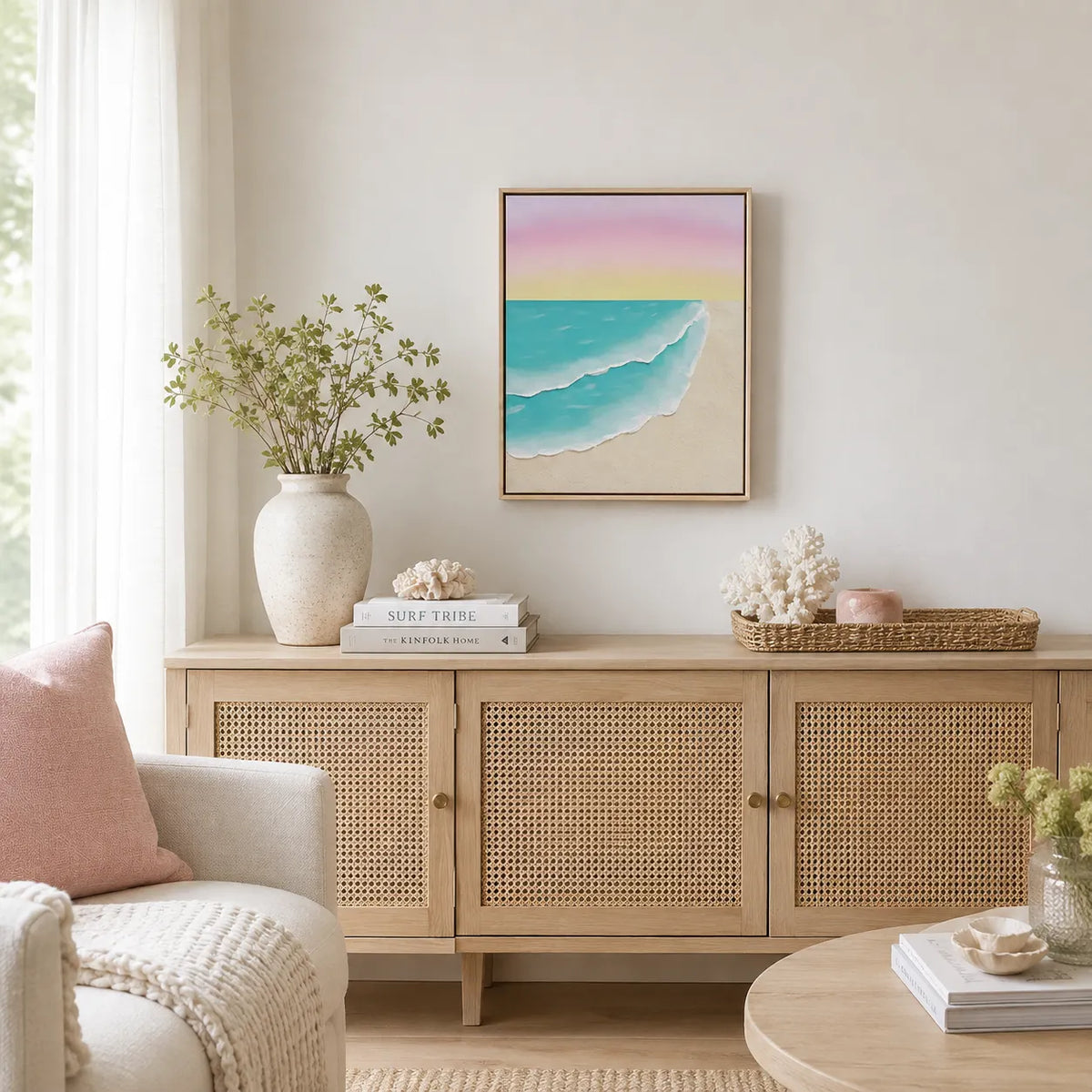 A framed pastel beach sunset painting hangs on a light beige wall above a wooden credenza in a bright, modern living room. The painting features a soft pink and yellow sky, turquoise ocean waves, and a sandy beige shore.