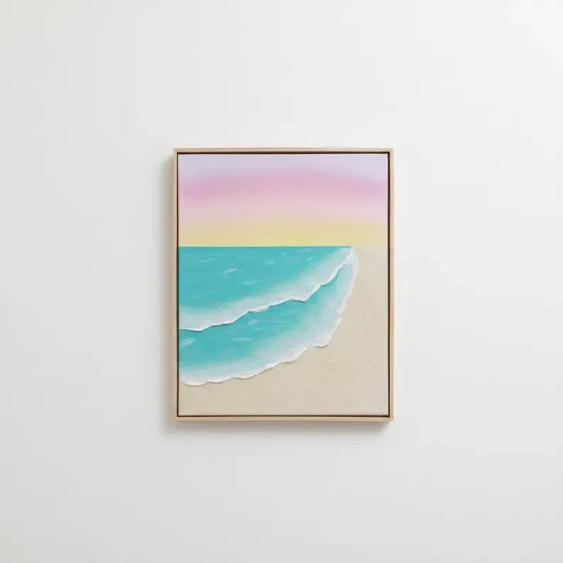 A framed minimalist beach art print featuring pastel pink and yellow sky, turquoise ocean waves, and a sandy beige shore, hung on a clean white wall.