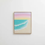A framed minimalist beach art print featuring pastel pink and yellow sky, turquoise ocean waves, and a sandy beige shore, hung on a clean white wall.