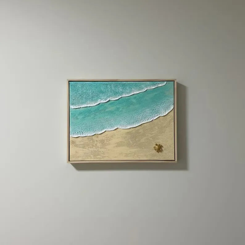 A framed square wall art print depicting turquoise ocean waves with white foam and a sandy beach, hung on a light gray wall.