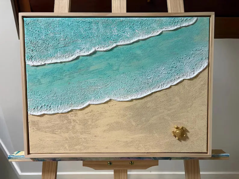 A framed beach scene artwork on an easel, showing turquoise ocean waves with white foam washing onto a sandy beige shore. The artwork appears textured and is photographed indoors against a light neutral wall.