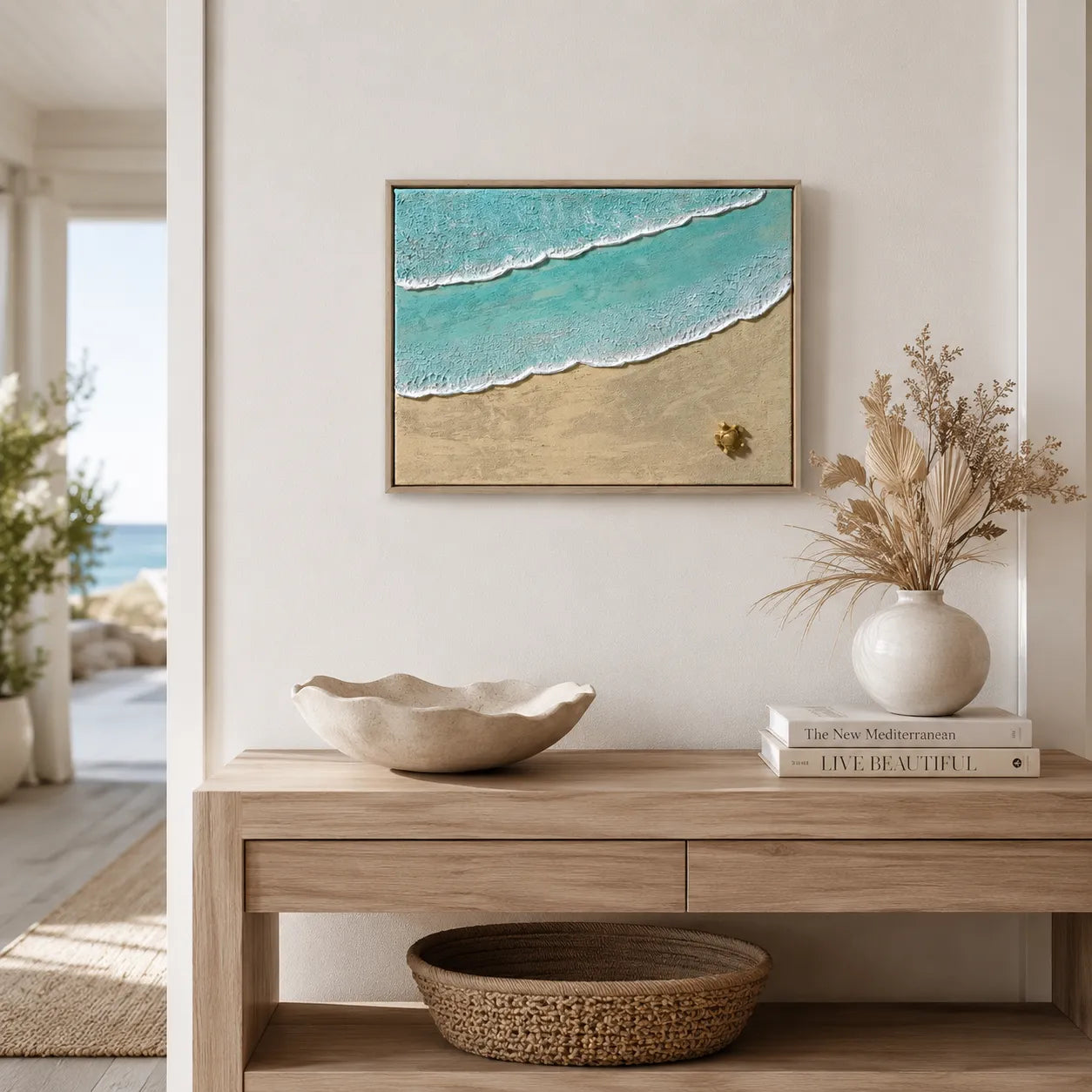 Framed wall art showing turquoise ocean waves rolling onto a sandy beach, displayed above a light wood console table in a bright, modern interior with decorative objects including a ceramic bowl, books, and a vase with dried grass.