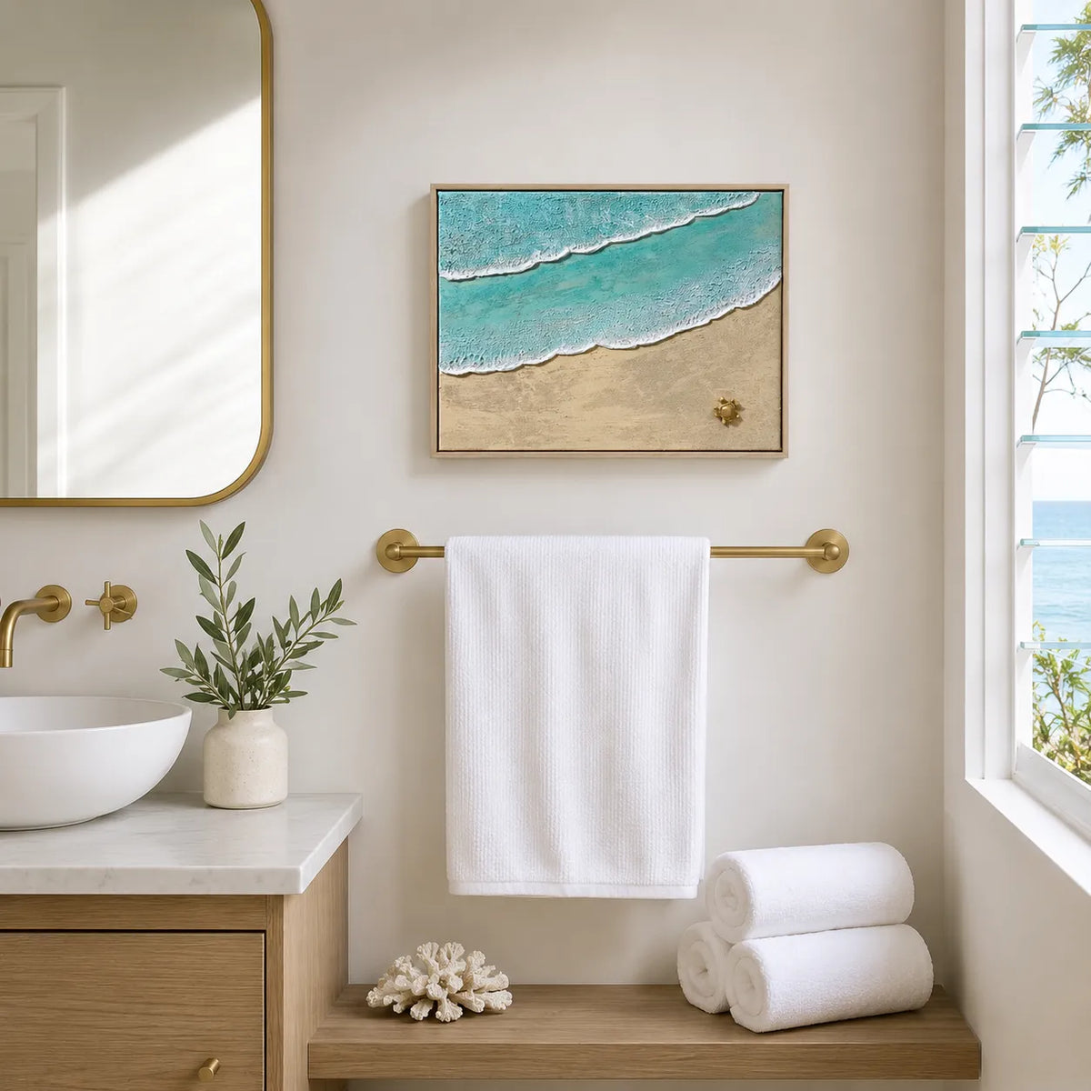 A framed beach wave art print hangs on a cream bathroom wall above a gold towel bar, with a white towel draped below. The print shows turquoise ocean water with white foam and sandy beige shore, styled in a bright, modern bathroom with a sink, mirror, and window.