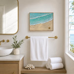 A framed beach wave art print hangs on a cream bathroom wall above a gold towel bar, with a white towel draped below. The print shows turquoise ocean water with white foam and sandy beige shore, styled in a bright, modern bathroom with a sink, mirror, and window.