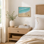 A framed beach art print with turquoise ocean waves and sandy shore, displayed on a cream wall above a light wood nightstand in a styled bedroom scene.