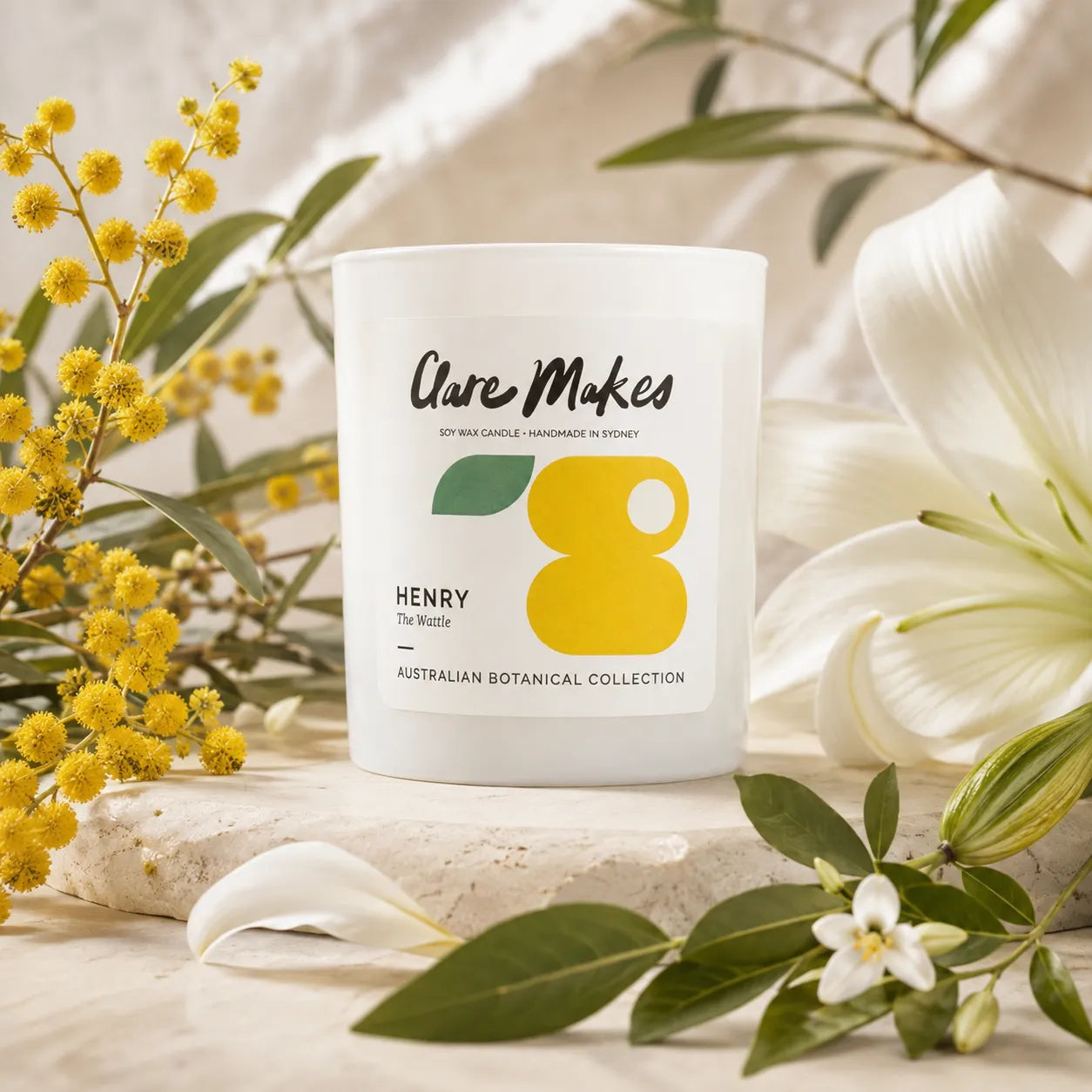 A white scented candle jar labeled 'Care Makes' and 'Henry' from their Australian Botanical Collection, styled with yellow mimosa flowers, a white lily, and green leaves on a stone slab.