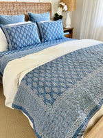 Blue patterned quilt and pillows on a bed in a bedroom setting.
