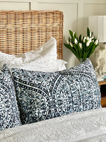 Decorative pillows on a couch with a wicker chair and lamp in the background