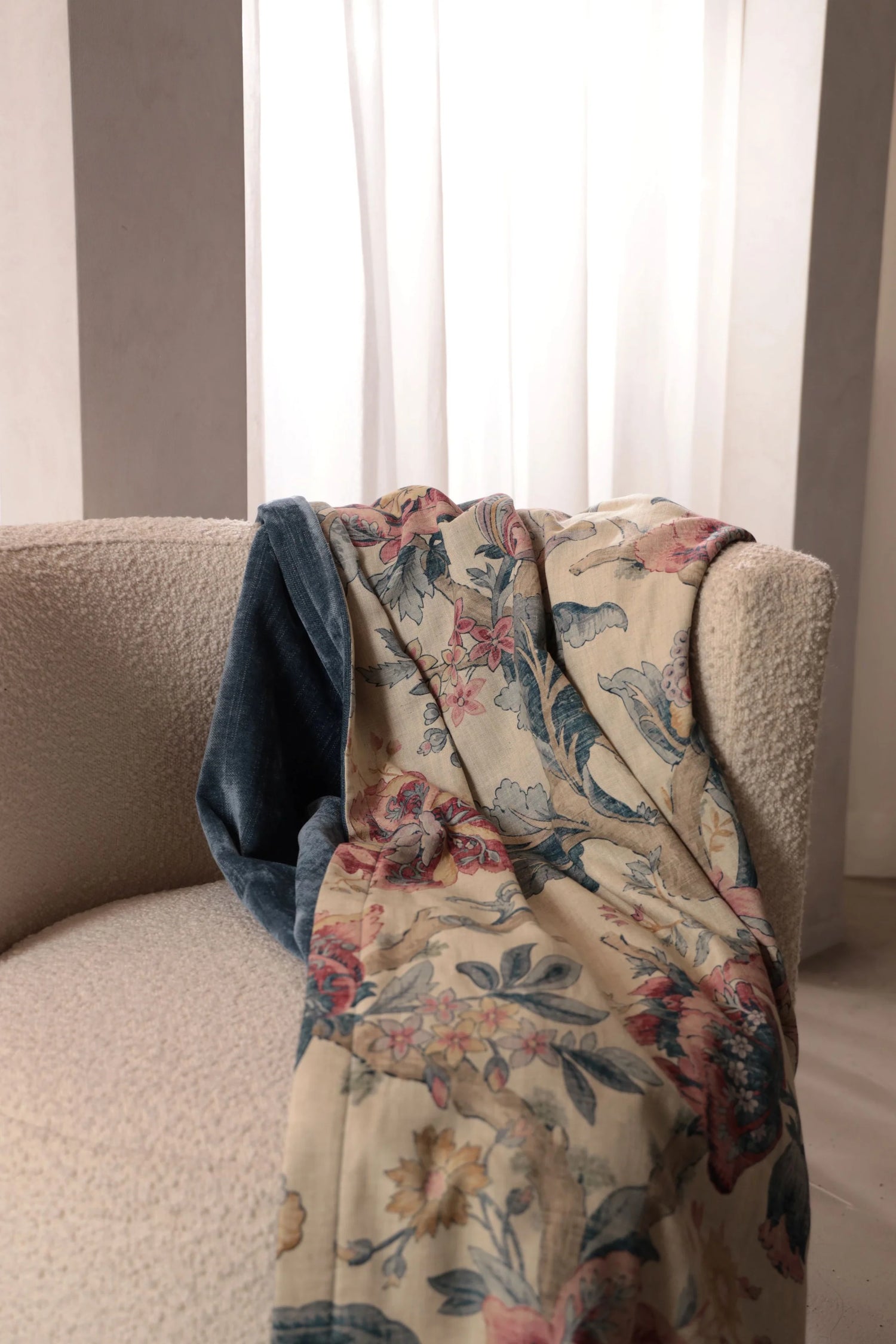 Floral-patterned blanket draped over a beige sofa with white curtains in the background
