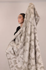 Woman wearing a large beige floral-patterned blanket over her shoulders against a plain background