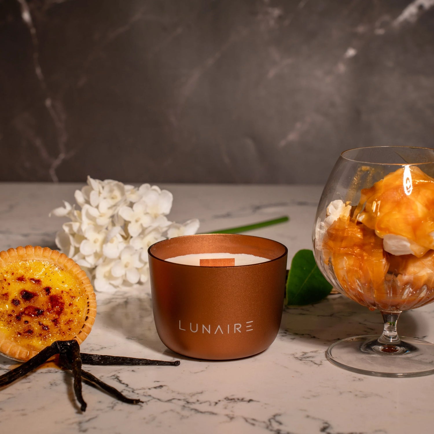 Candle with 'LUNAIRE' branding on a marble surface with vanilla beans and flowers.