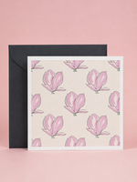 A square greeting card with a repeating pink magnolia floral pattern on a warm cream background, shown in front of a black envelope against a soft pink backdrop.