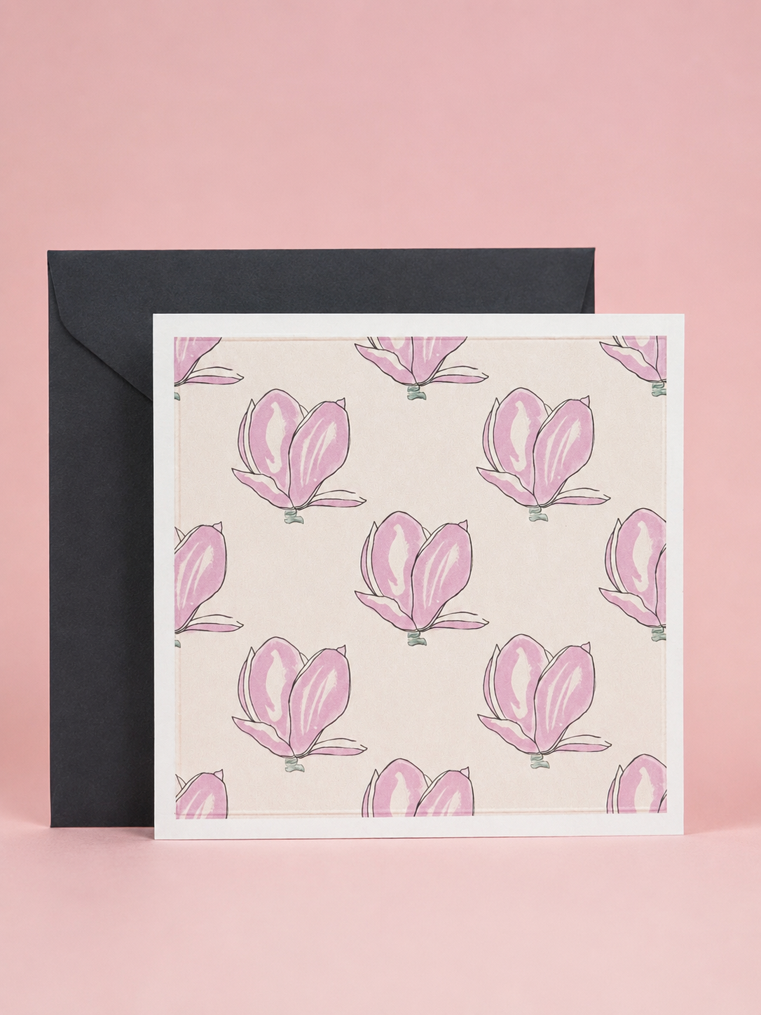 A square greeting card with a repeating pink magnolia floral pattern on a warm cream background, shown in front of a black envelope against a soft pink backdrop.