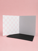 An open greeting card with a pink and white polka dot pattern on the left panel, a plain light gray center panel, and a black envelope placed in front, set against a solid pink background.
