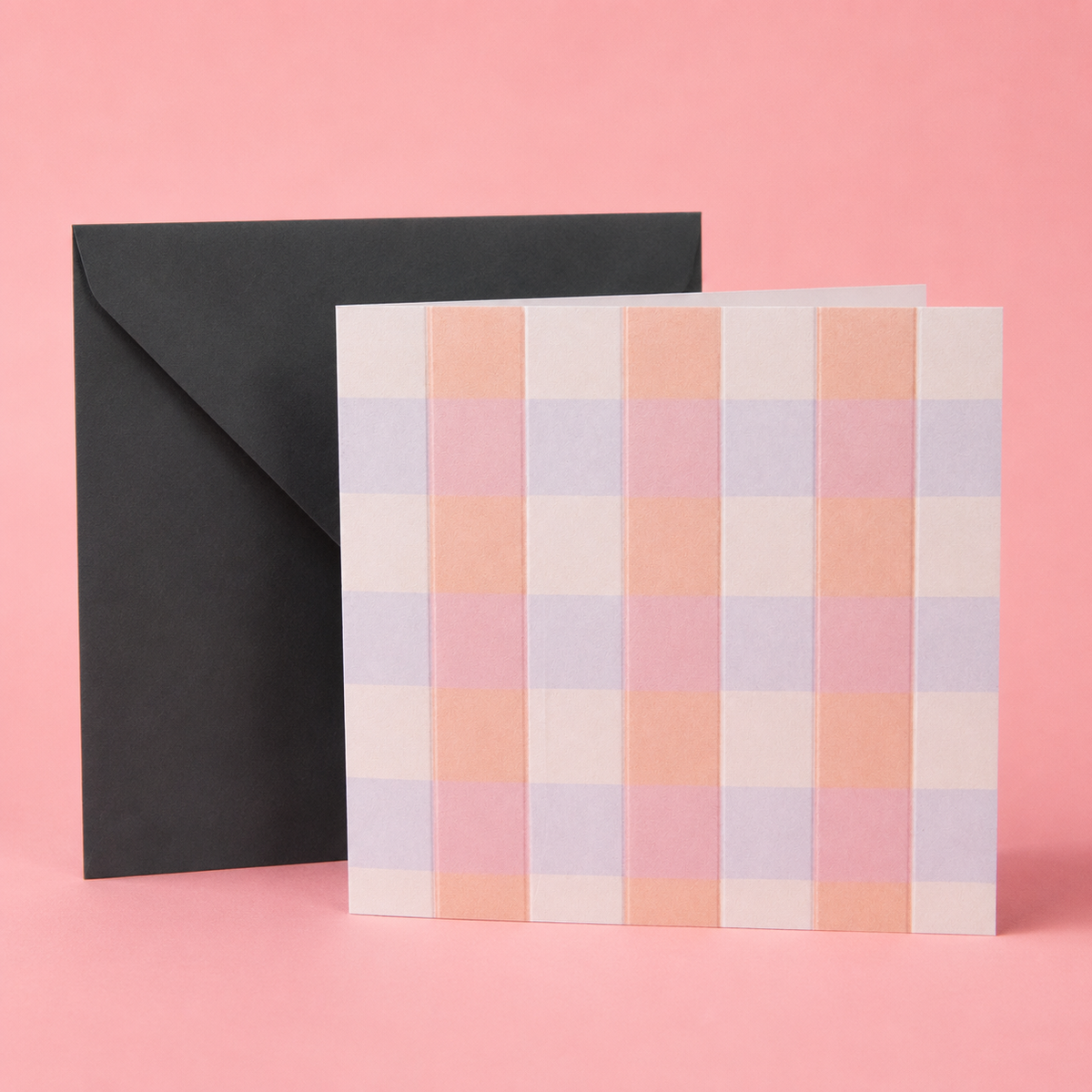 A square greeting card with a pastel plaid pattern in pink, peach, lavender, and white, shown standing in front of a black envelope on a solid pink background.