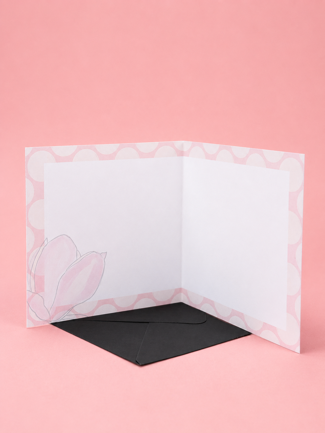 An open white greeting card with a pink patterned border and a light pink tulip illustration on the lower left, displayed with a black envelope on a solid pink background.