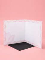 An open white greeting card with a pink patterned border and a light pink tulip illustration on the lower left, displayed with a black envelope on a solid pink background.
