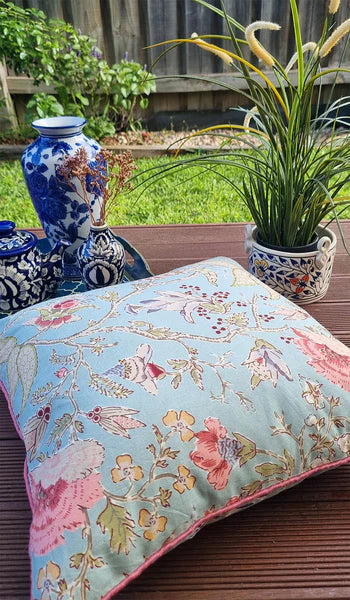 Floral-patterned cushion on a wooden surface with decorative vases and plants in the background.