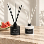 Two reed diffusers on a stone countertop: one matte black bottle with black reeds and one white bottle with white reeds, both labeled Luna Blu London Sunday Lunch Watermelon. Sliced watermelon sits to the left, with warm natural light and leaf shadows in the background.