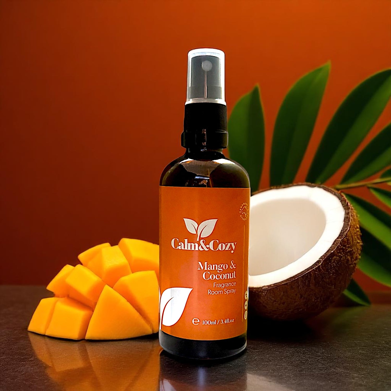 Bottle of Calm & Cozy Mango & Coconut room spray with mango pieces and a coconut on a reflective surface.