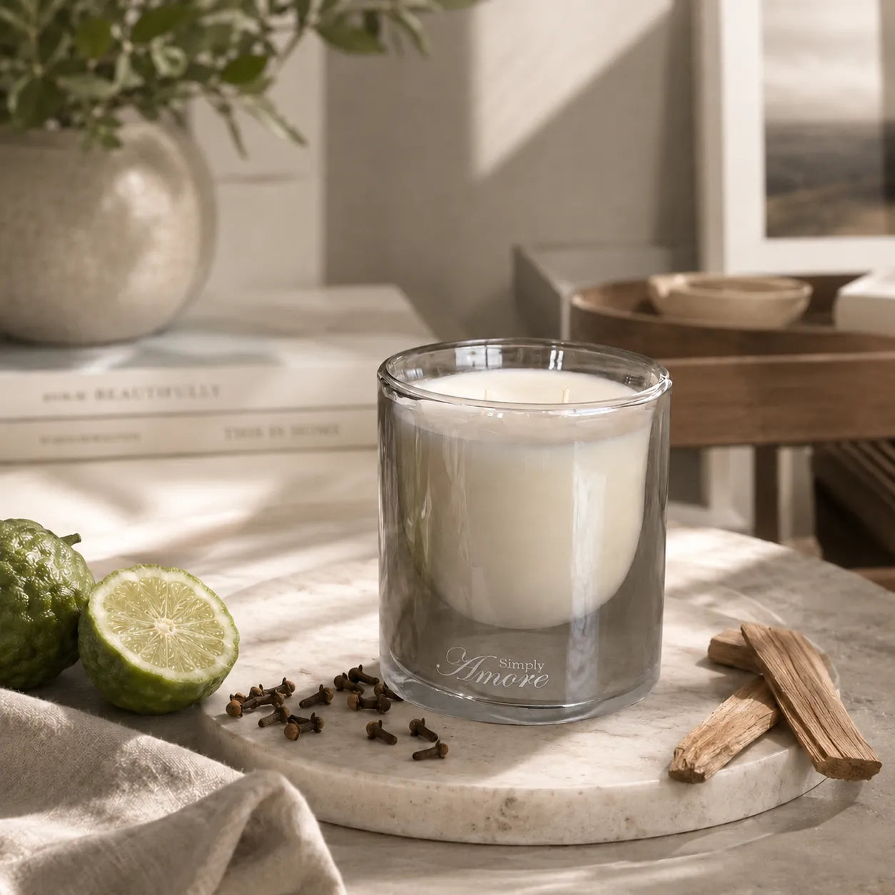 A clear glass candle jar filled with white wax sits on a round marble tray, surrounded by a green lime, cloves, and wood pieces in a softly styled indoor setting.