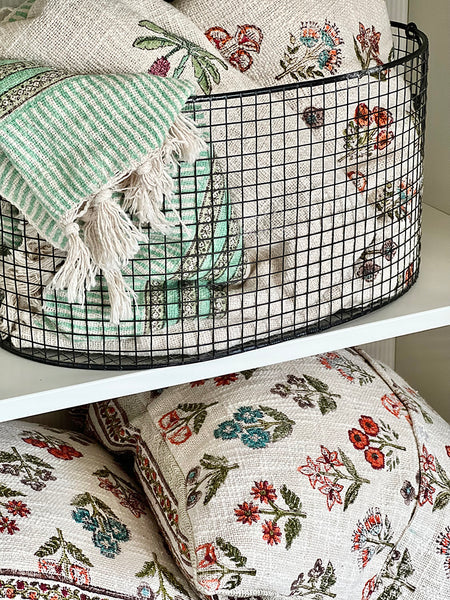 Decorative pillows with floral patterns and a green and white striped towel in a wire basket.