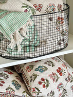 Decorative pillows with floral patterns and a green and white striped towel in a wire basket.