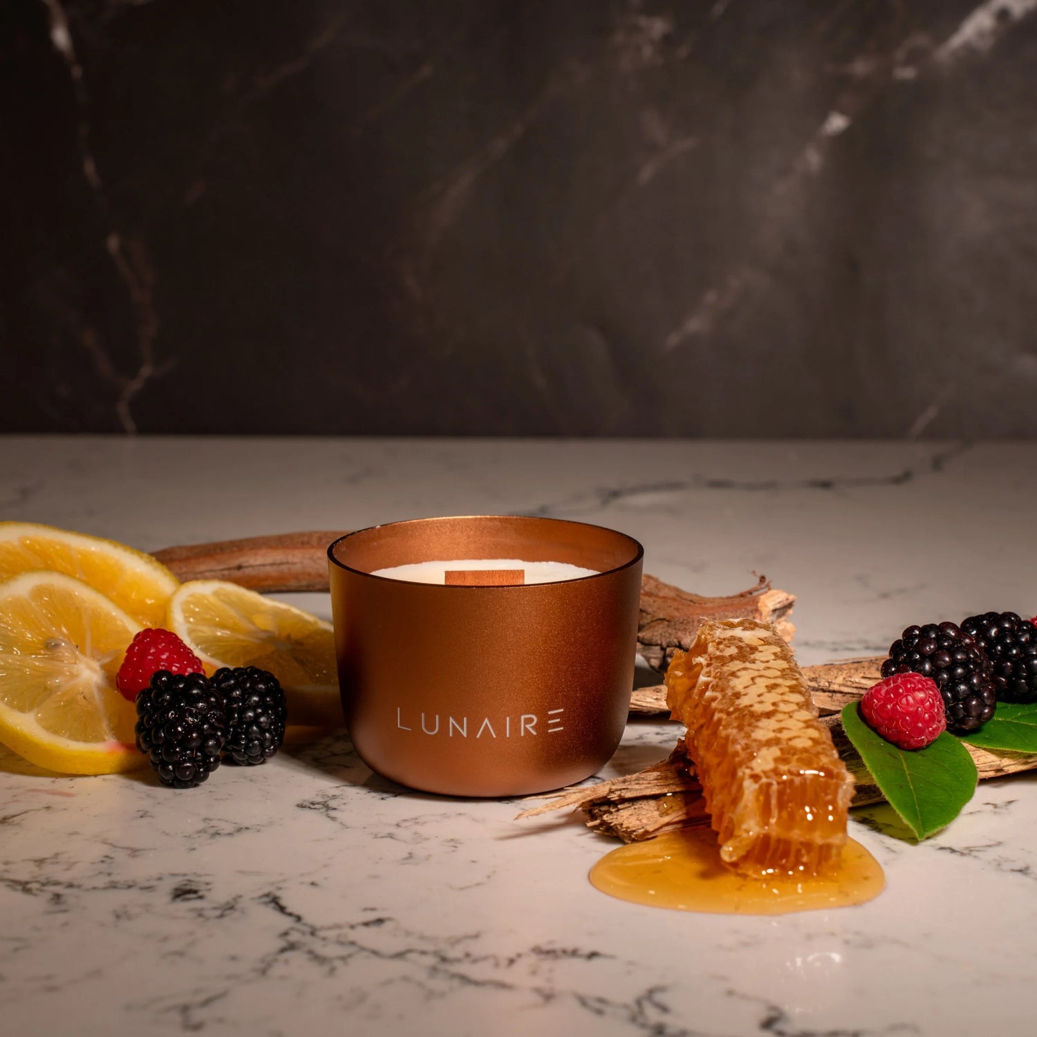 Bronze 'LUNAIRE' candle with honeycomb, lemons, and berries on a marble surface