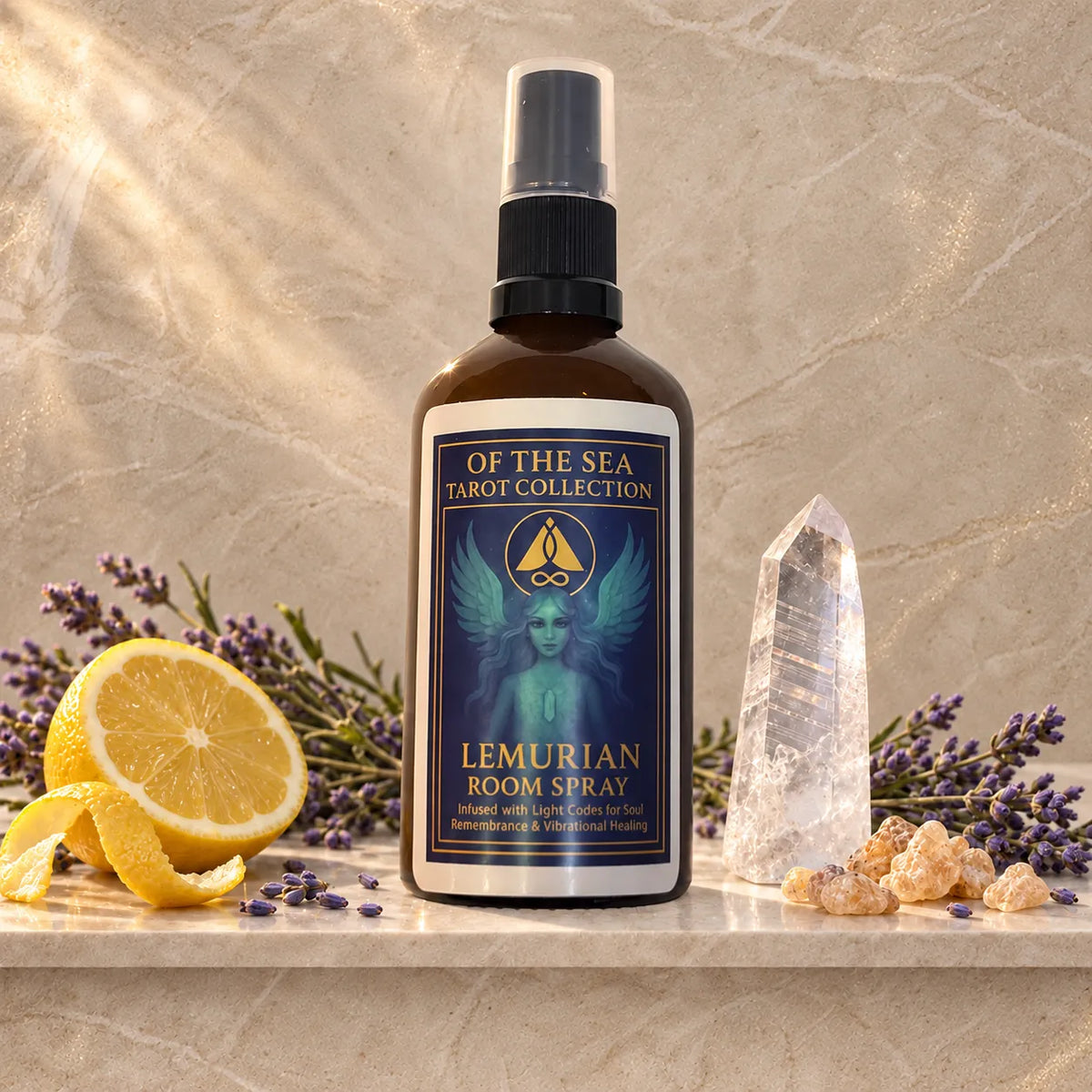 A bottle of Of The Sea Tarot Collection Lemurian Room Spray stands on a stone surface with lemon slices, lavender sprigs, a clear crystal tower, and small dried flowers around it. The bottle is amber-brown glass with a black spray top and a dark blue label featuring a winged figure and gold text.