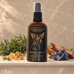 A bottle of Archangel Michael Room Spray from the 'Of the Sea Tarot Collection' displayed on a marble shelf with natural props including a crystal, rosemary, and wood shavings against a textured stone background.