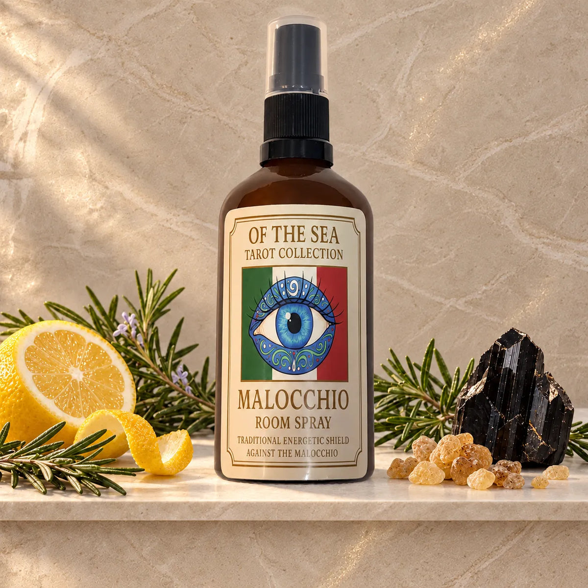 A bottle of Of The Sea Tarot Collection Malocchio Room Spray stands on a stone surface with lemon slices, rosemary sprigs, and a black crystal cluster around it. The bottle is amber brown with a black spray top and a cream label featuring a blue eye over the Italian flag colors.