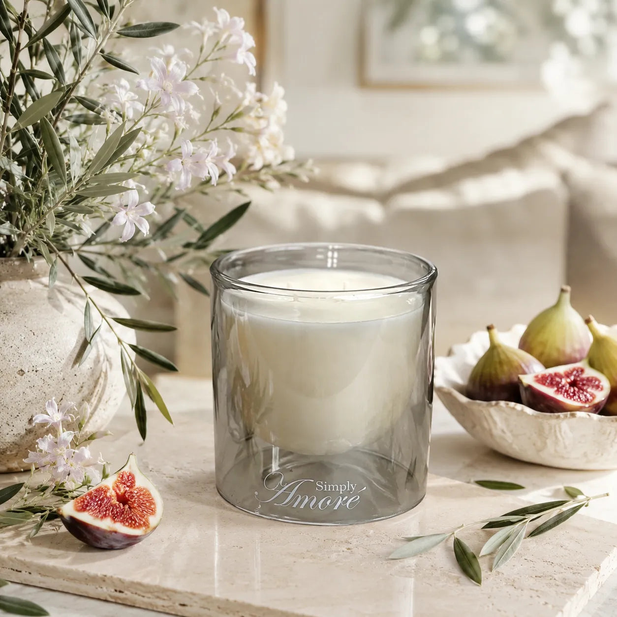 A frosted gray glass jar candle with white wax sits on a light stone surface, styled with figs, olive branches, and white flowers in a softly lit indoor setting. The candle is branded 'Simply A more'.