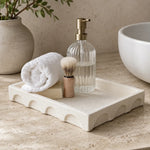 A rectangular cream stone bathroom tray with scalloped edges, styled on a beige stone countertop with a rolled white towel, soap bar, shaving brush, ribbed clear soap dispenser, and part of a white vessel bowl in the background.