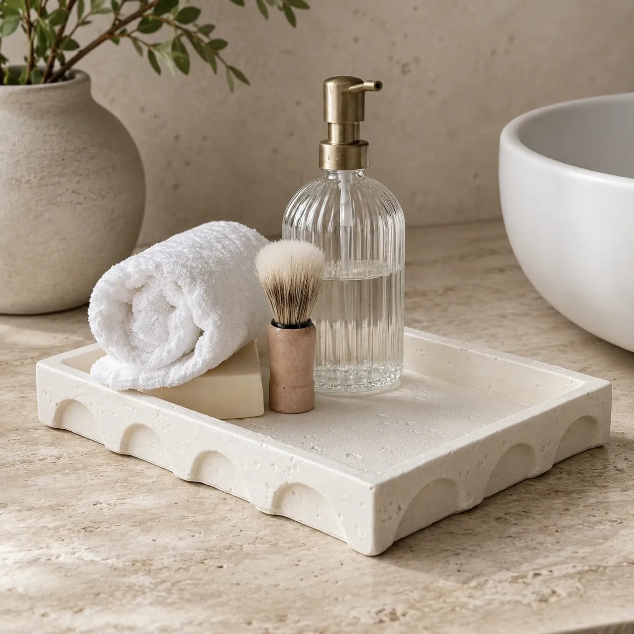 A rectangular cream stone bathroom tray with scalloped edges, styled on a beige stone countertop with a rolled white towel, soap bar, shaving brush, ribbed clear soap dispenser, and part of a white vessel bowl in the background.
