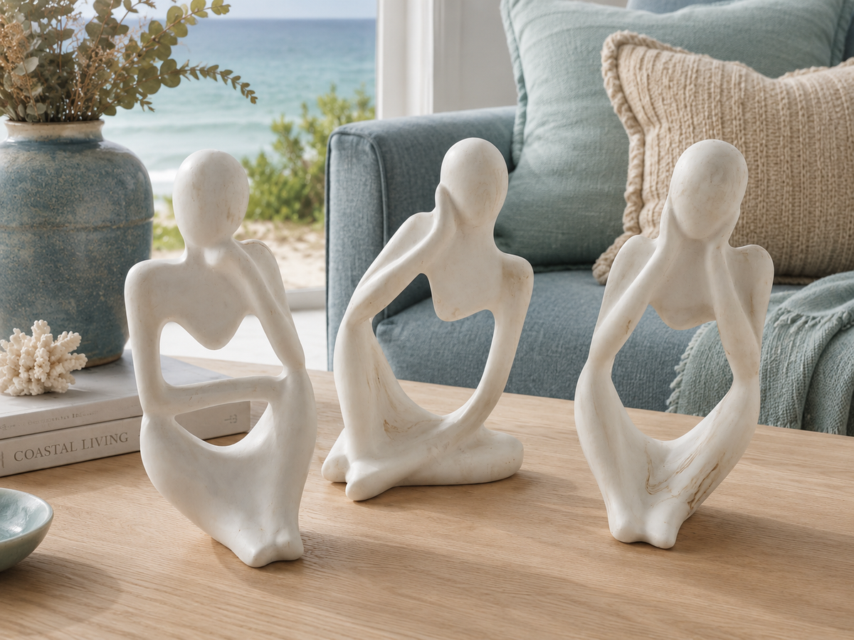Three white ceramic figurines of seated abstract human figures arranged on a light wood table in a living room setting, with a blue sofa, pillows, and a vase with flowers in the background.